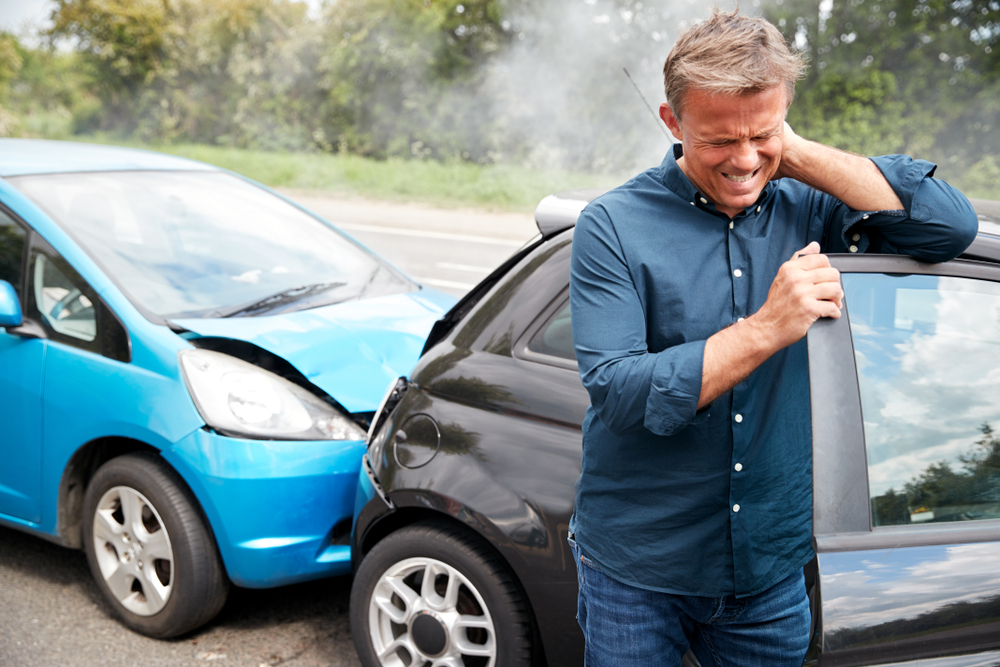 Man in blue button down shirt standing next to car holding neck after being rear-ended