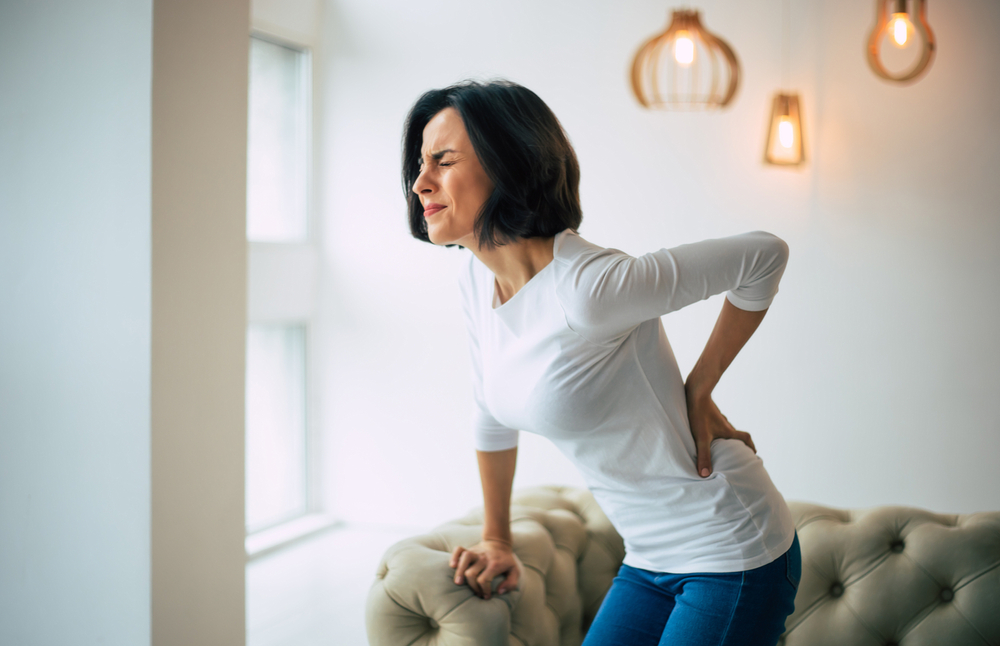 Adult woman is holding her lower back, while standing and suffering from unbearable pain.