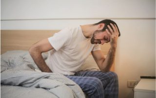 man sits on edge of bed, head in hands, tired