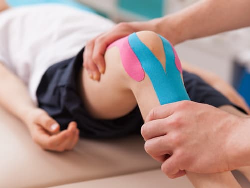 A healthcare professional applies colorful kinesiology tape to a patient’s knee during a physiotherapy or sports injury rehabilitation session, providing support and promoting recovery.