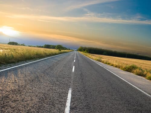 A long, empty paved road stretches through open countryside fields under a colorful sunrise sky, symbolizing travel, opportunity, freedom, and the journey ahead.