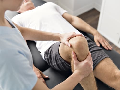 A physiotherapist gently assesses a patient’s bent knee during a rehabilitation or physiotherapy session, with the patient lying on a treatment table in a clinical setting.
