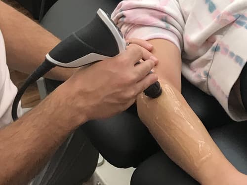 A healthcare professional administers shockwave therapy to a patient’s lower leg using a handheld device during a rehabilitation session, helping promote healing, reduce pain, and support recovery from tendon and soft tissue injuries.