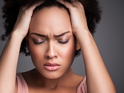 Close-up of a woman holding her head with both hands and displaying signs of discomfort, illustrating headache, migraine pain, stress, tension, or mental fatigue.