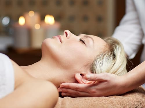 A woman lying comfortably on a massage table while a therapist supports her head and neck during a relaxing spa or wellness treatment in a calm setting.